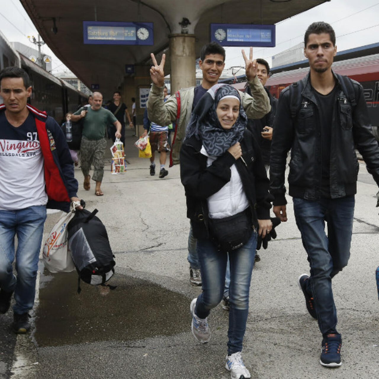 Migranten mit Gepaeck auf einem Bahnsteig, im Hintergrund Anzeigetafeln mit Zugzielen