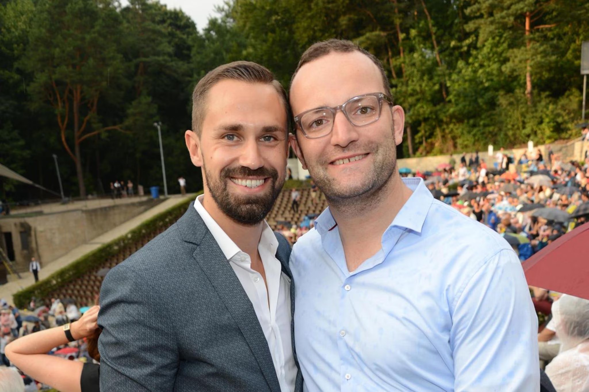 Jens Spahn und sein Ehemann Daniel Funke stehen eng nebeneinander bei einer Open-Air-Veranstaltung und laecheln in die Kamera, im Hintergrund ein gefuelltes Zuschauerrund