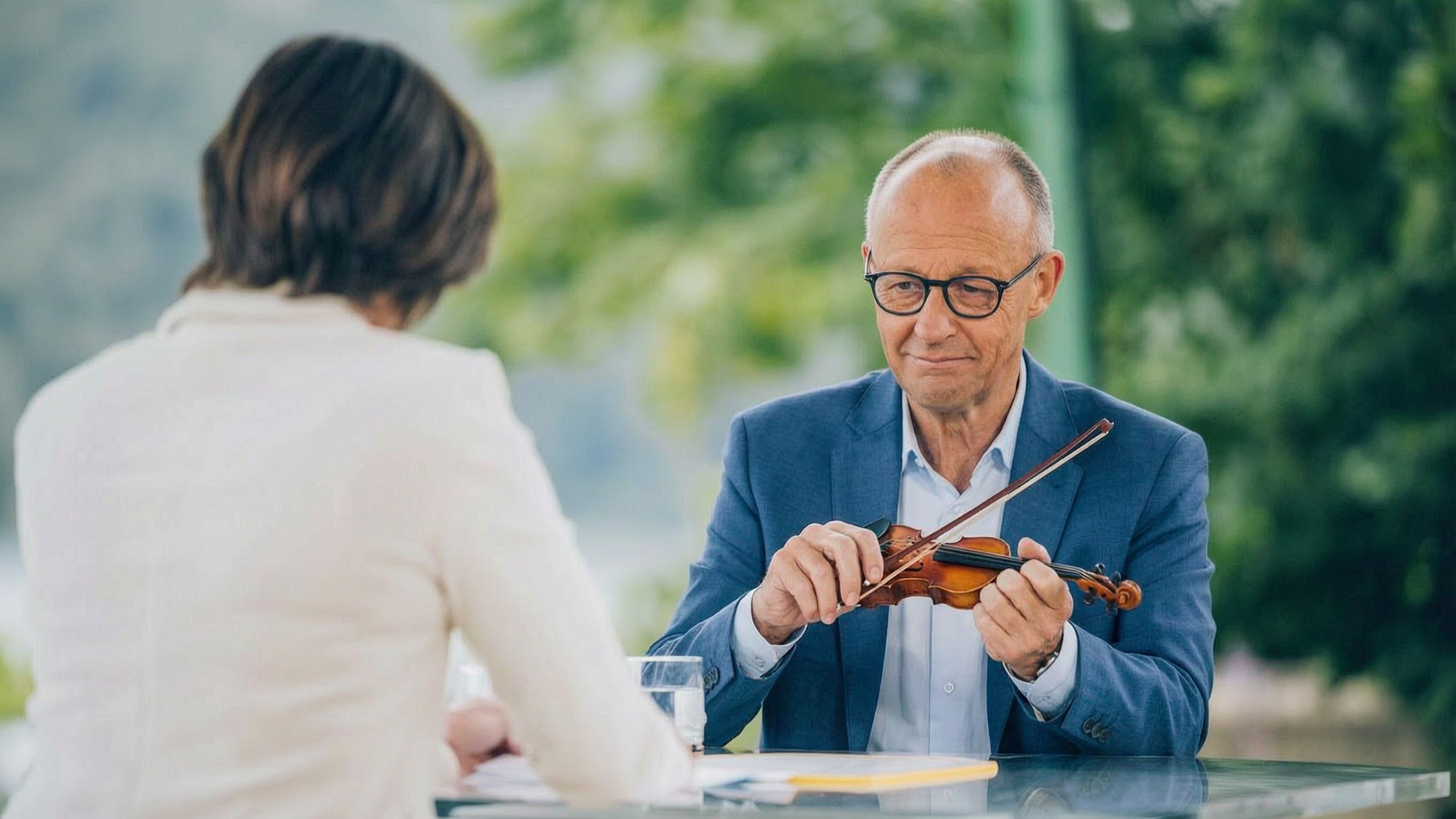 Bundeskanzler Friedrich Merz sitzt im sommerlichen ZDF-Interview am Tisch und spielt eine winzige Geige, ihm gegenueber eine Interviewerin von hinten