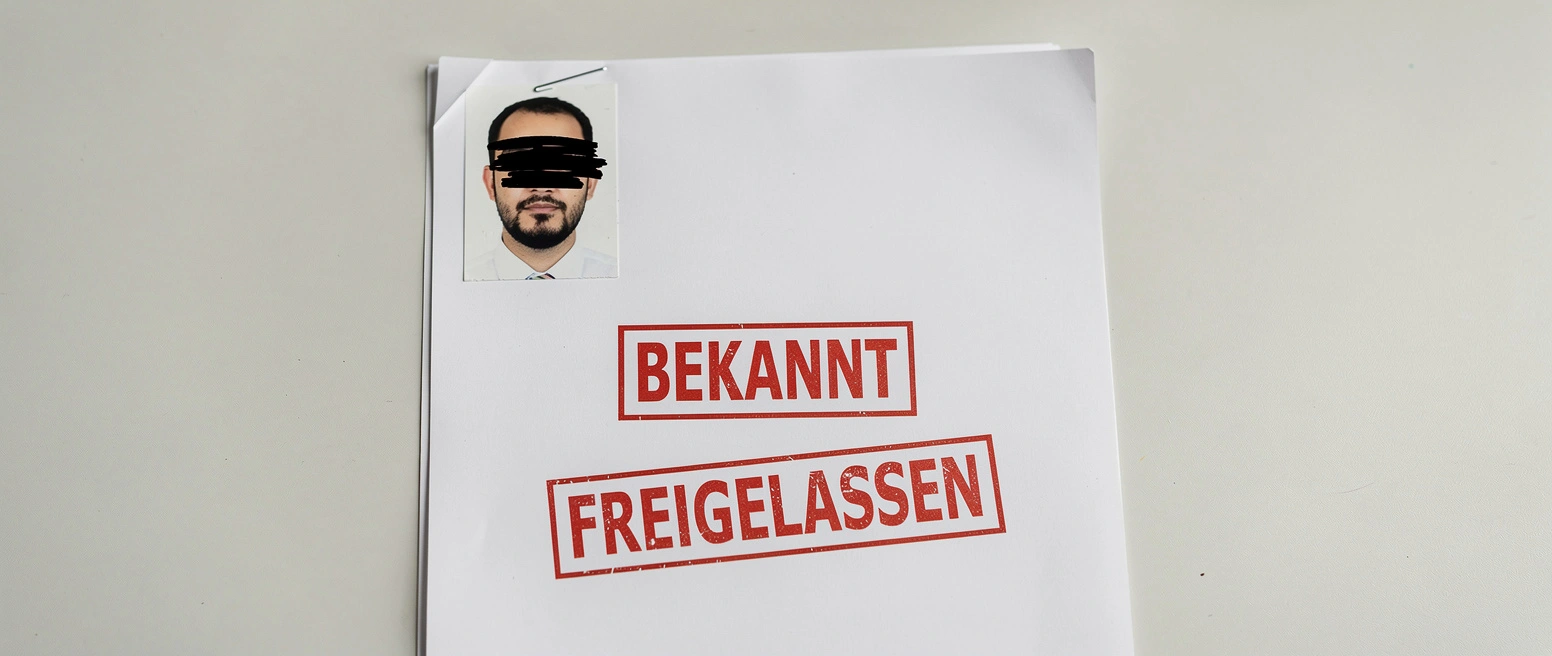 Eine geoeffnete Ermittlungsakte auf einem Tisch mit zwei roten Stempeln BEKANNT und FREIGELASSEN und einem angeklammerten geschwaerzten Passfoto