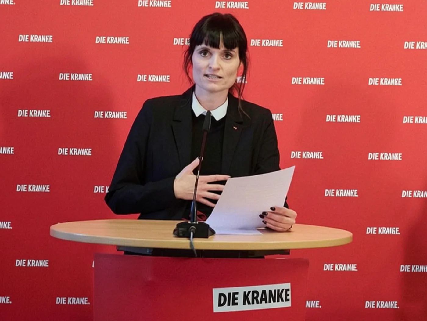 Satirische Fotomontage: Politikerin am Rednerpult vor roter Parteiwand, auf der statt des Parteinamens ueberall Die Kranke steht