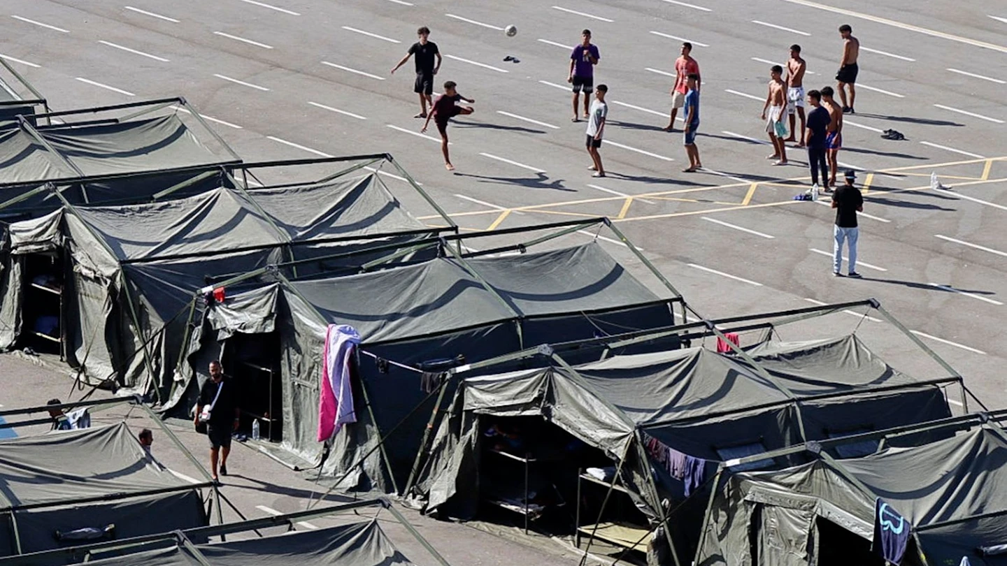 Provisorisches Zeltlager auf einer asphaltierten Fläche in Ceuta; hinter den Zelten spielen junge Männer Fußball