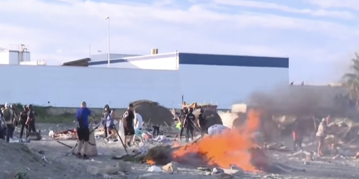 Menschen in einem provisorischen Lagerbereich in Ceuta; im Vordergrund brennt ein offenes Feuer